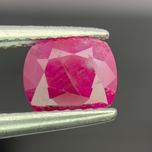 May include: A cushion-cut vibrant pink gemstone held by metal tweezers. The stone features a faceted surface with visible internal inclusions and a deep, saturated color profile that reflects light across its geometric, polished top facets.