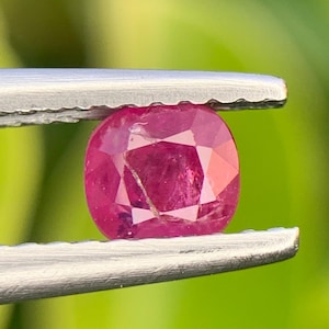 May include: A vibrant, cushion-cut ruby gemstone with a deep pinkish-red hue and visible internal facets. The gemstone is held securely between the serrated metal tips of a pair of silver-toned tweezers, showcasing its polished surface.