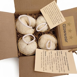 Pode incluir: Uma caixa de papelão contendo quatro esponjas de luffa redondas, bege e fibrosas, com alças de corda branca. A caixa inclui um cartão marrom com o texto Glowwear e três estrelas, e um segundo cartão com instruções de uso das esponjas.