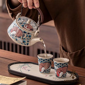 Puede incluir: Una tetera de cerámica y dos tazas a juego decoradas con carpas koi rojas y patrones de olas azules. La tetera cuenta con un asa trenzada de color marrón oscuro y una tapa. El conjunto se dispone sobre una bandeja rectangular con el texto HIKARYCLAY.