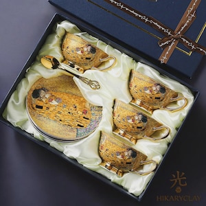 Könnte beinhalten: Ein Teeservice in einer dunkelblauen Geschenkbox mit Satinfutter, bestehend aus drei goldfarbenen Keramiktassen und einer passenden Untertasse, alle mit einer Reproduktion des Gemäldes Der Kuss verziert. Ein goldfarbener Metalllöffel liegt bei.