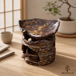 May include: A dark brown ceramic oil burner with a textured, bark-like exterior and a wide, shallow bowl top. The base features a circular opening for a tea light candle. The text HIKARYCLAY and a Japanese kanji character are printed in the corner.