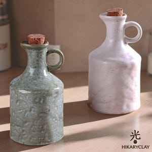 May include: Two ceramic oil cruets with cork stoppers, one in a speckled sage green glaze and the other in a speckled off-white glaze. Each bottle features a side handle, a narrow neck, and a pouring spout. The text Hikaryclay is visible.
