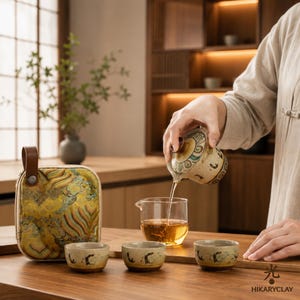 May include: A ceramic tea set featuring a patterned teapot with a lid, three small matching tea cups, and a clear glass pitcher containing amber tea. A square, yellow-patterned fabric carrying case with a brown leather handle sits nearby.