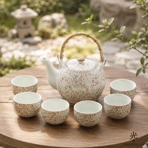 Puede incluir: Un juego de té de cerámica que consta de una tetera redonda con asa de bambú y seis tazas de té pequeñas a juego. La tetera y las tazas presentan un delicado patrón floral con flores rosas y marrones y enredaderas verdes sobre fondo blanco.
