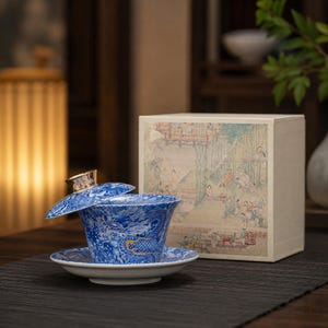 Puede incluir: Un juego de té gaiwan de porcelana azul y blanca con un motivo de dragón detallado y detalles dorados. El set incluye un cuenco, un platillo y una tapa con un pomo dorado, acompañado de una caja cuadrada decorada con una ilustración paisajista.