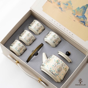 May include: A tea set in a beige gift box featuring four small cups and a teapot with a lid, all decorated with a gold, teal, and white floral pattern. The box lid displays a landscape painting and the text BEAUTIFUL MOUNTAINS AND RIVERS.