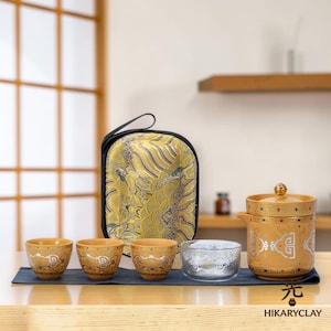 以下が含まれることがあります： 銀色の模様が入った3つの小さな黄色の陶器製カップ、透明なガラスボウル、蓋付きの黄色の陶器製ティーポットで構成されたティーセット。カップの後ろには黄色の柄入りキャリングケースが置かれ、すべて紺色のランナーの上に配置されています。