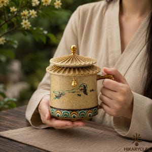 Pode incluir: Uma caneca de cerâmica com acabamento bege texturizado, apresentando uma tampa em forma de telhado com um remate dourado e um pequeno pendente dourado. A caneca exibe uma cena pintada de figuras em movimento e uma borda decorativa de ondas.