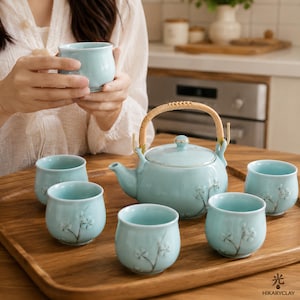 Puede incluir: Un juego de té de cerámica azul claro que consta de una tetera con asa de ratán curvada y cinco tazas de té pequeñas a juego. Cada pieza presenta un delicado diseño de rama floral pintado a mano en blanco y marrón sobre una bandeja de madera.