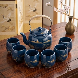 May include: A dark blue ceramic tea set consisting of a teapot with a woven handle and five matching cups. Each piece features a gold-colored landscape design depicting mountains and trees. The teapot has a lid and a curved spout.