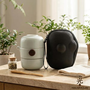May include: A light green ceramic tea set consisting of a stacked teapot and cup, featuring a dark brown circular wooden accent. The set includes a small glass jar with dried tea leaves, a wooden scoop, and a black protective carrying case.