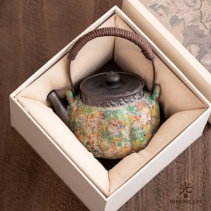 Puede incluir: Una tetera de cerámica con un acabado texturizado y moteado multicolor en tonos verde, amarillo y rojo. Presenta una tapa gris oscuro, un pico vertedor gris oscuro y un asa curva envuelta en cordón marrón dentro de una caja blanca.
