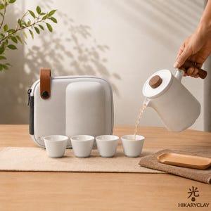Puede incluir: Una tetera de cerámica blanca con tapa y asa de madera vertiendo té en una de las cuatro tazas de cerámica blanca a juego. Un estuche protector gris con una correa de cuero marrón se encuentra detrás de las tazas y una bandeja de madera.