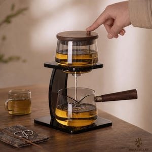 May include: A glass tea brewing set featuring a top chamber with a dark wood lid and a lower glass pitcher with a matching dark wood handle. The set is mounted on a black stand, with tea flowing from the top chamber into the pitcher below.