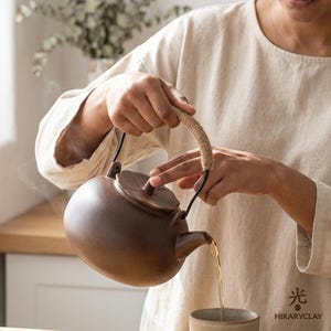 May include: A dark brown matte ceramic teapot with a rounded body, a lid shaped like a leaf, and a curved metal handle wrapped in light tan rope. The teapot is pouring steaming tea into a matching ceramic cup. The text HIKARYCLAY is visible.