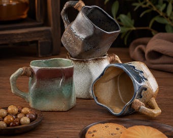 May include: Three ceramic mugs with irregular, faceted shapes and textured surfaces. One mug features a dark grey glaze, another a speckled white finish, and the third a blend of green and red tones. Each handle is wrapped with natural twine.