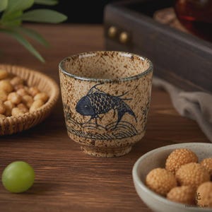 Könnte beinhalten: Eine Keramik-Teetasse mit gesprenkelter beiger Glasur und einer dunkelblauen, handgemalten Fischillustration, die über stilisierten Wellen schwimmt. Die Tasse hat einen konischen Boden und einen breiten Rand mit einer rustikalen, texturierten Oberfläche.