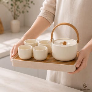 May include: A cream-coloured ceramic tea set featuring a cylindrical teapot with a curved wooden handle and a matching lid with a small wooden knob, accompanied by three small matching tea cups, all arranged on a rectangular tray.