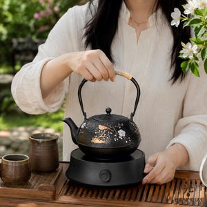 May include: A black cast iron teapot with a textured surface, featuring gold and white painted floral and bird motifs. The teapot has a curved handle wrapped in light-coloured material, a matching lid, and a spout, resting on a black electric base.