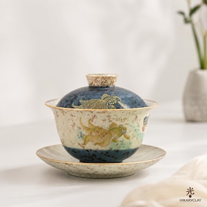 Może przedstawiać: Ceramiczny zestaw do herbaty gaiwan składający się z miseczki, pokrywki i spodka z nakrapianą kremowo-niebieską glazurą. Zewnętrzna strona ozdobiona jest złotymi ilustracjami złotych rybek. Pokrywka posiada teksturowany uchwyt.