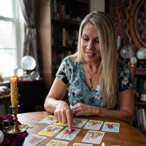 Puede incluir: Una persona con cabello rubio largo que viste una camiseta tie-dye azul y blanca y un collar con colgante de cristal, se&ntilde;alando una carta de tarot de tres de espadas sobre una mesa de madera con una vela amarilla encendida en un candelabro.