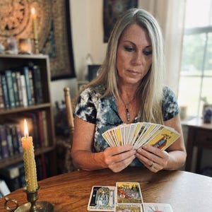 Op de afbeelding: Een waaier van tarotkaarten, met vier extra tarotkaarten uitgespreid op een houten tafel. E&eacute;n kaart is gelabeld met "High Priestess". Een hoge, getextureerde gele kaars in een koperen kandelaar staat aan de linkerkant van de kaarten.