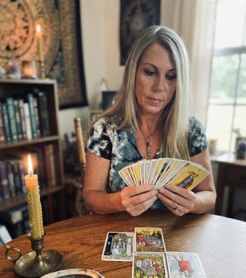 Op de afbeelding: Een waaier van tarotkaarten, met vier extra tarotkaarten uitgespreid op een houten tafel. E&eacute;n kaart is gelabeld met "High Priestess". Een hoge, getextureerde gele kaars in een koperen kandelaar staat aan de linkerkant van de kaarten.