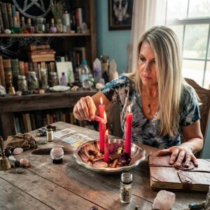 Puede incluir: Un cuenco de metal plateado que contiene tres velas de pilar rojas encendidas, rodeado de varios cristales, una botella de vidrio peque&ntilde;a, cartas de tarot, un libro encuadernado en cuero, una esfera de cristal y una peque&ntilde;a campana de lat&oacute;n.