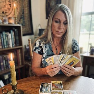 Puede incluir: Una persona sostiene un abanico de cartas del tarot con una baraja extendida en sus manos. Sobre la mesa de madera frente a ella hay cuatro cartas del tarot dispuestas, incluida una etiquetada como The High Priestess. Una vela amarilla encendida.