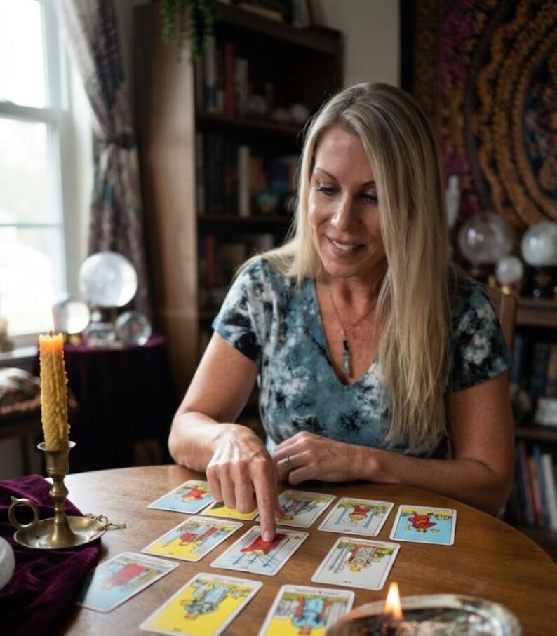Op de afbeelding: Een persoon met lang blond haar in een blauw en wit tie-dye shirt zit aan een ronde houten tafel. Ze wijzen naar een tarotkaart uit een reeks kaarten die op de tafel ligt. Een hoge gele bijenwaskaars in een koperen houder staat erbij.