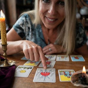 Puede incluir: Un conjunto de cartas de tarot extendidas sobre una mesa de madera, destacando la carta del Tres de Espadas en el centro. La carta muestra un coraz&oacute;n rojo atravesado por tres espadas azules sobre un fondo blanco. Una vela amarilla encendida est&aacute; cerca.