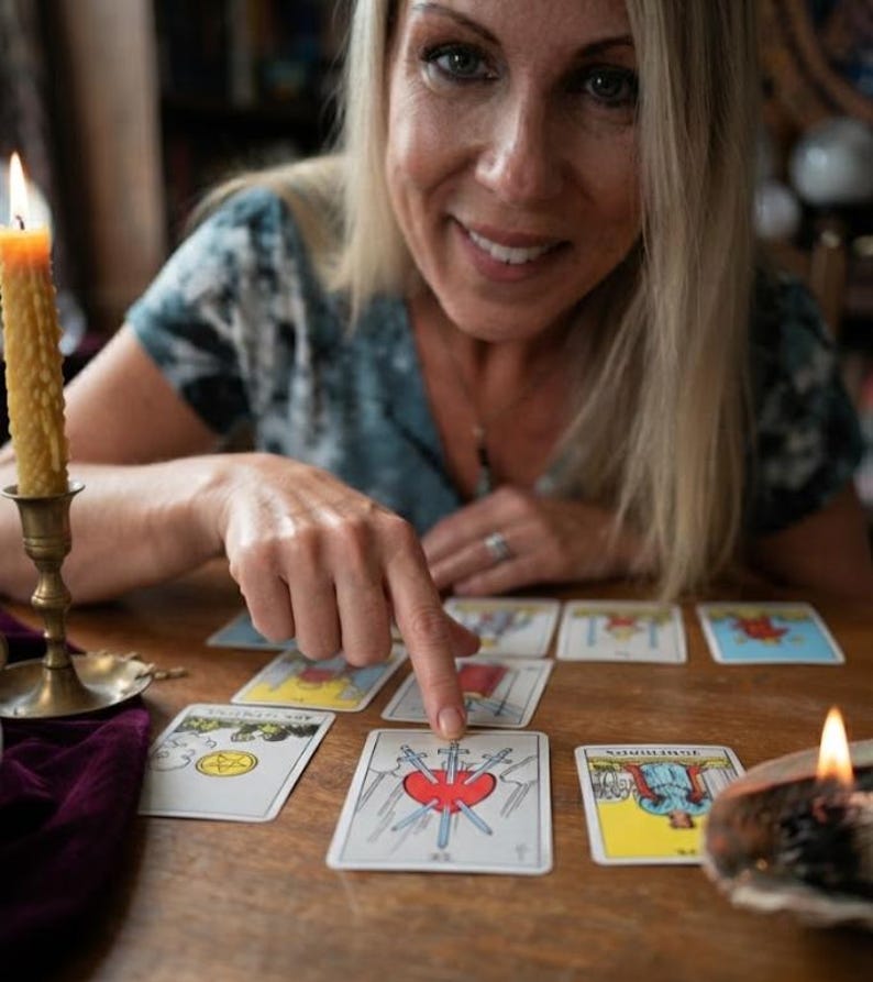 Puede incluir: Un conjunto de cartas de tarot extendidas sobre una mesa de madera, destacando la carta del Tres de Espadas en el centro. La carta muestra un coraz&oacute;n rojo atravesado por tres espadas azules sobre un fondo blanco. Una vela amarilla encendida est&aacute; cerca.