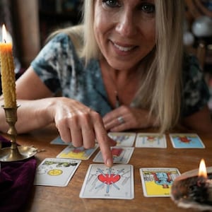 Op de afbeelding: Een set tarotkaarten verspreid over een houten tafel, met de kaart Drie van Zwaarden prominent in beeld met een rood hart doorboord door drie blauwe zwaarden, naast andere ge&iuml;llustreerde kaarten en twee brandende bijenwaskaarsen in houders.