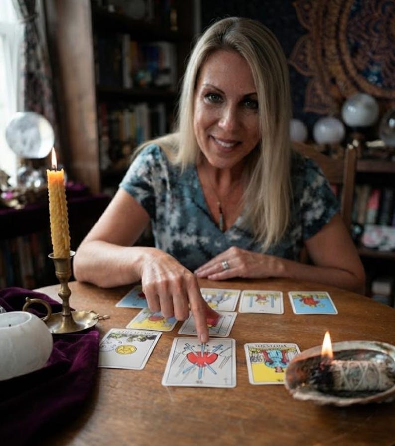 Op de afbeelding: Een set tarotkaarten verspreid over een houten tafel, met de Drie van Zwaarden kaart prominent in het midden. Een brandende bijenwaskaars in een koperen houder en een brandende saliebundel in een abalone schelp liggen op de tafel bij de kaarten.