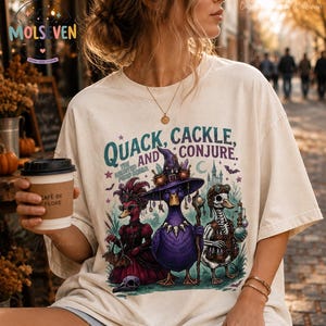 Könnte beinhalten: Ein cremefarbenes T-Shirt mit einem Grafikdruck und dem Text "QUACK, CACKLE, AND CONJURE. THE HAUNTED POND SQUAD." Die Illustration zeigt drei Enten, die als Hexe, Vampir und Skelett verkleidet sind, umgeben von verschiedenen magischen Elementen.