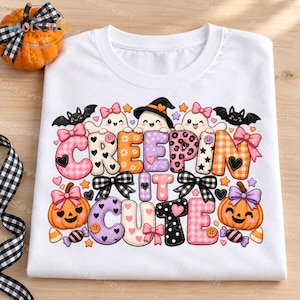 Puede incluir: Una camiseta blanca con un gráfico colorido y el texto "CREEPIN IT CUTE". El diseño incluye fantasmas, calabazas, murciélagos y estrellas de dibujos animados en tonos rosa, naranja, morado y negro, con lazos y varios patrones.