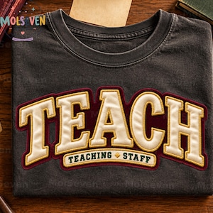 May include: A charcoal gray crew neck t-shirt featuring the word TEACH in large, cream-colored, embroidered varsity-style letters with a dark red outline. Below, a smaller banner reads TEACHING STAFF in dark green text with a gold diamond.