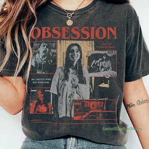 May include: A charcoal gray short-sleeve t-shirt featuring a vintage-style graphic print. The design includes the word OBSESSION in large red letters, multiple black and white film-style scenes, and the text BE CAREFUL WHO YOU WISH FOR.