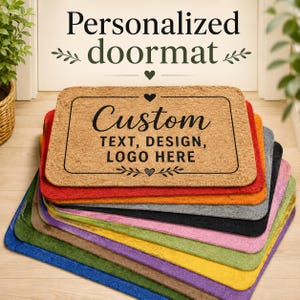 May include: A stack of rectangular doormats in various colors including tan, red, orange, gray, black, pink, yellow, green, and blue. The top tan mat features black text that reads Custom TEXT, DESIGN, LOGO HERE with heart and leaf accents.