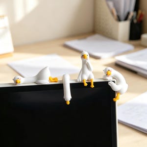 Könnte beinhalten: Ein Set aus vier weißen Gänsefiguren mit orangefarbenen Schnäbeln und Füßen, die so gestaltet sind, dass sie auf der Oberkante eines Computermonitors sitzen. Jede Figur zeigt eine andere Pose: liegend, hängend, aufrecht sitzend und angelehnt.