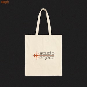 May include: A cream-colored canvas tote bag with two shoulder straps. The front of the bag features a graphic logo in orange and dark brown, displaying the text "studio reject" alongside a stylized crosshair icon with a central diamond shape.