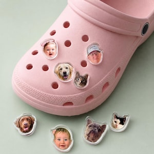 May include: A light pink foam clog shoe featuring several clear, custom photo charms inserted into the ventilation holes. The charms display portraits of various people, a golden retriever, a brown dog, a French bulldog, and two cats.