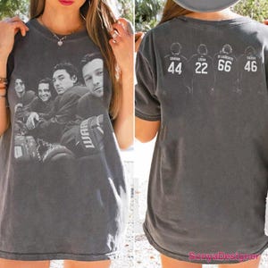 May include: A charcoal gray short-sleeve t-shirt featuring a black and white photographic print of four individuals on the front. The back displays four outlined figures with the names Graham 44, Logan 22, Di Laurentis 66, and Tucker 46 printed above.