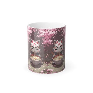 May include: A white ceramic mug featuring a repeating illustration of a grey cat wearing a white and red fox mask, eating a bowl of ramen with chopsticks under a pink cherry blossom tree with falling petals scattered on the ground below.