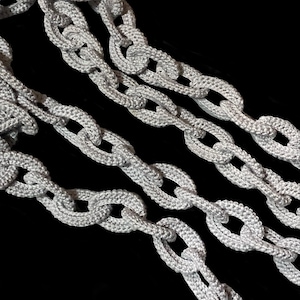 May include: A decorative chain composed of interlocking oval links crafted from a light grey, textured, woven fabric material. The chain features a repeating pattern of connected loops that create a flexible, rope-like structure with a consistent finish.