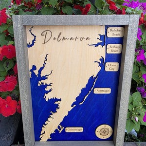 Könnte beinhalten: Eine gerahmte Holzkarte der Delmarva-Halbinsel mit einer hellen Holzlandmasse vor einem tiefblauen Wasserhintergrund. Die Karte enthält eingravierte Textbeschriftungen für Rehoboth Beach, Bethany Beach, Ocean City, Assateague und Chincoteague.