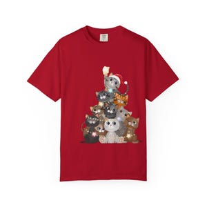 May include: A red short-sleeved t-shirt featuring a graphic print of a stack of cartoon cats arranged in a triangular shape. The cats are various colors, including gray, brown, and orange, with some wearing Santa hats and wrapped in string lights.
