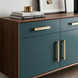Op de afbeelding: Een donkerblauwgroen dressoir met een frame van walnoothout en goudkleurige metalen handgrepen. Het meubel heeft twee lades en twee kastdeuren. Bovenop liggen twee gestapelde boeken met de titels design en DESIGNBOOK onder een gouden cilinder.