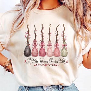 May include: A cream-colored t-shirt featuring a graphic of six stylized broomsticks decorated with pink bows and leaves. Below the brooms, the text reads A Wise Woman Chooses Well in a cursive font, accented by small pink floral and leaf illustrations.